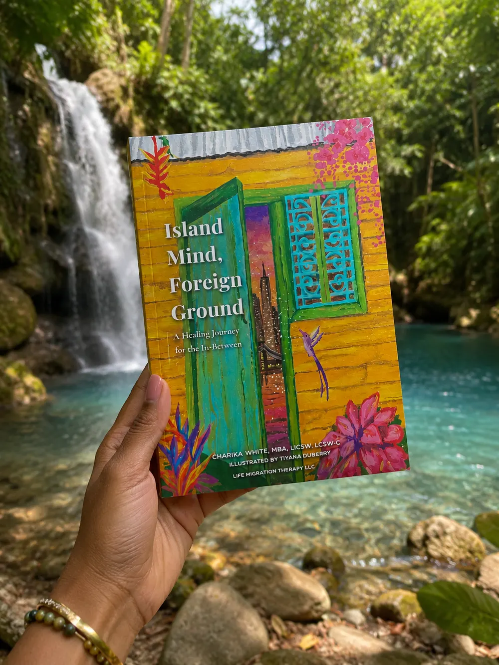 A hand holding Island Mind, Foreign Ground up in front of a waterfall and a turquoise pool, the cover painting bright against the green.