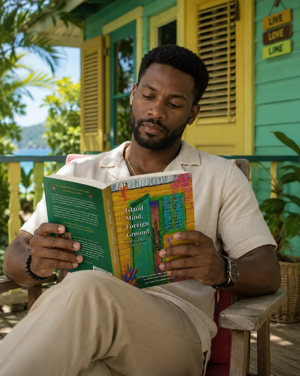 A man sitting on a painted Caribbean veranda reading Island Mind, Foreign Ground, holding it open with the sea behind him.