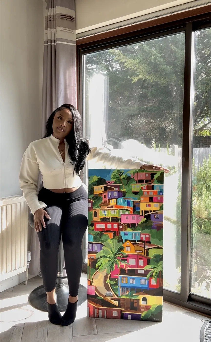 Tiyana Duberry standing beside one of her paintings, a hillside of brightly coloured Caribbean houses among palms and greenery.