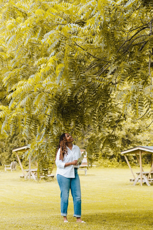 Charika White standing barefoot on grass under a broad walnut tree, looking up through the leaves, near picnic tables